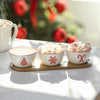 Three white bowls with red Christmas designs on a wooden tray, filled with marshmallows and candy, in front of a blurred Christmas tree.
