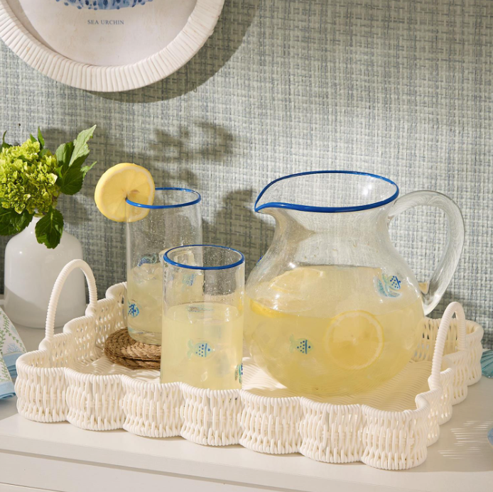 Clear glass pitcher and glasses with lemon slices on a white woven basket tray against a textured wall.