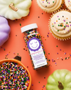 A bottle of Sweetapolita Halloween sprinkles displayed on a bright orange background with seasonal props including mini pumpkins, cupcakes, and a bowl of sprinkles.