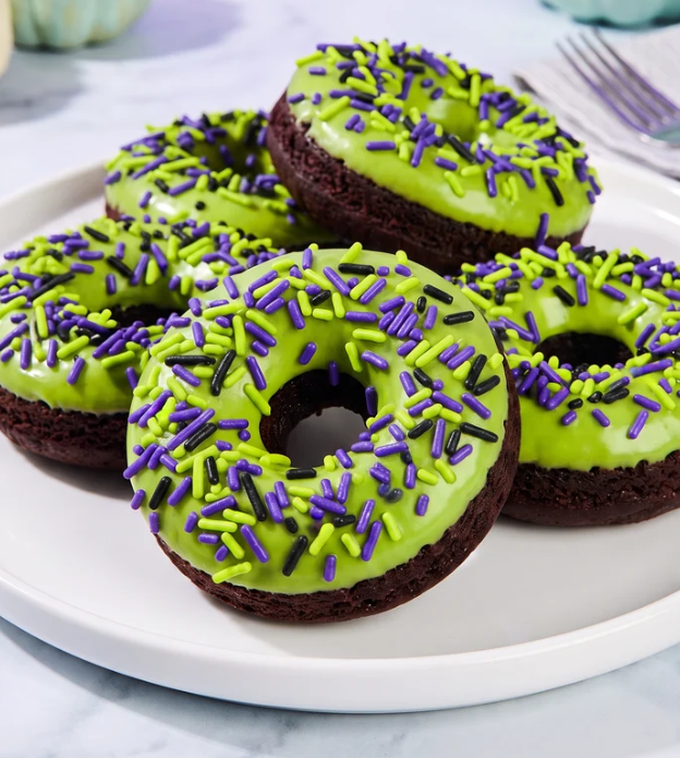 Four chocolate cake donuts with bright lime green frosting and colorful sprinkles, arranged on a white plate.
