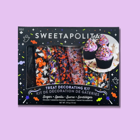 Sweetapolita Halloween treat decorating kit in a black box with colorful sprinkles, and decorated cupcakes visible through a clear window, set against a solid lavender background.