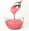 Pink frosting being poured from a spoon into a clear glass bowl on a white background