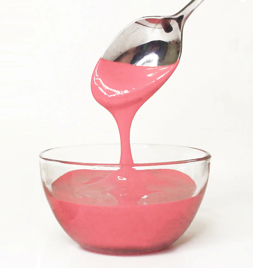 Pink frosting being poured from a spoon into a clear glass bowl on a white background