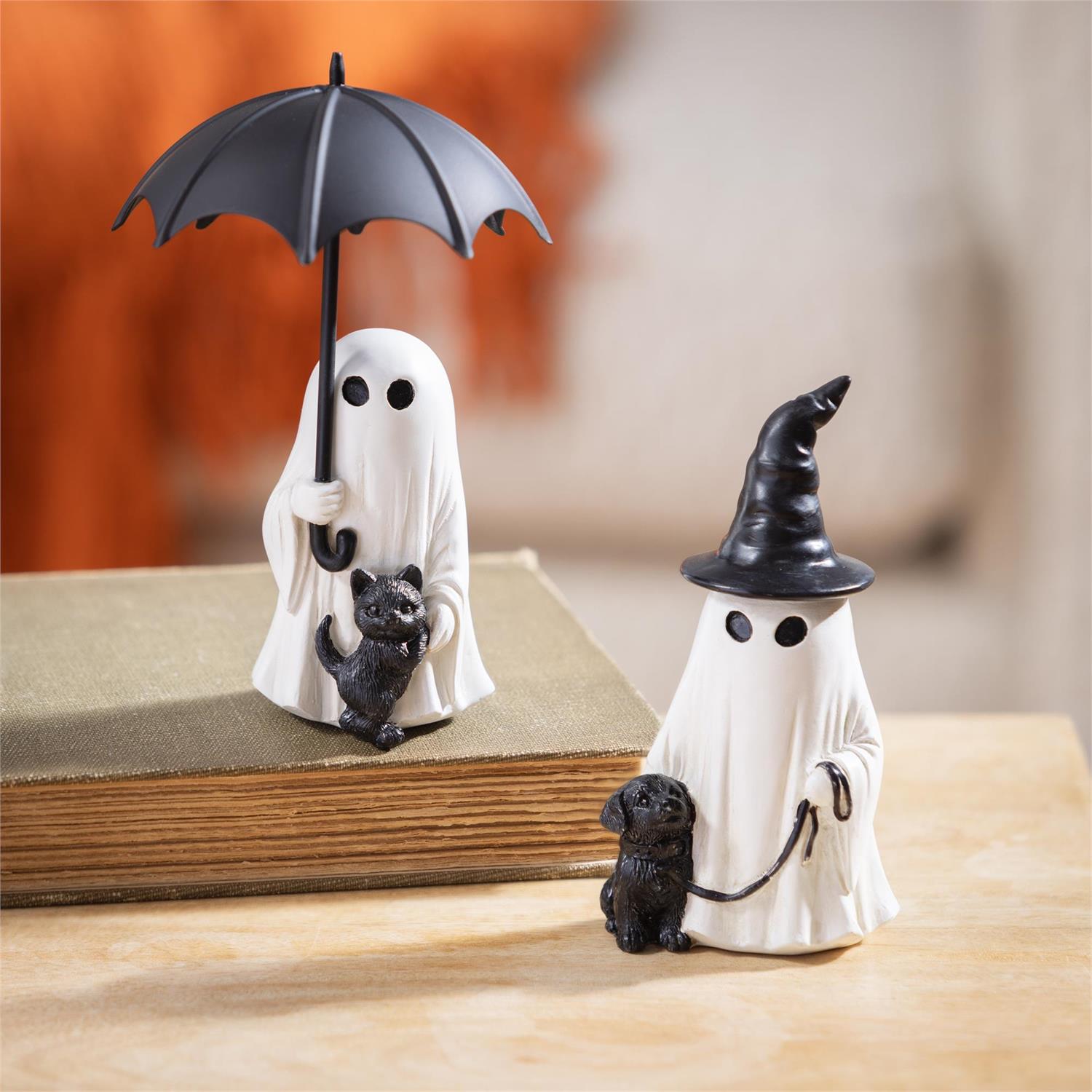 Two small figurines of ghosts with a black hat, umbrella and a cat and dog on a wooden surface.