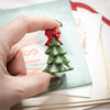 Hand holding a Christmas tree-shaped decoration with a red bow over a card with text.
