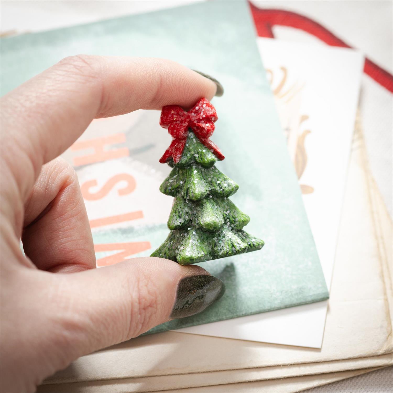 Hand holding a Christmas tree-shaped decoration with a red bow over a card with text.