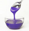 Spoon pouring purple frosting into a clear glass bowl on a white background