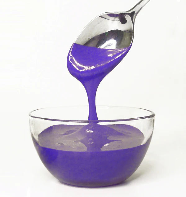 Spoon pouring purple frosting into a clear glass bowl on a white background