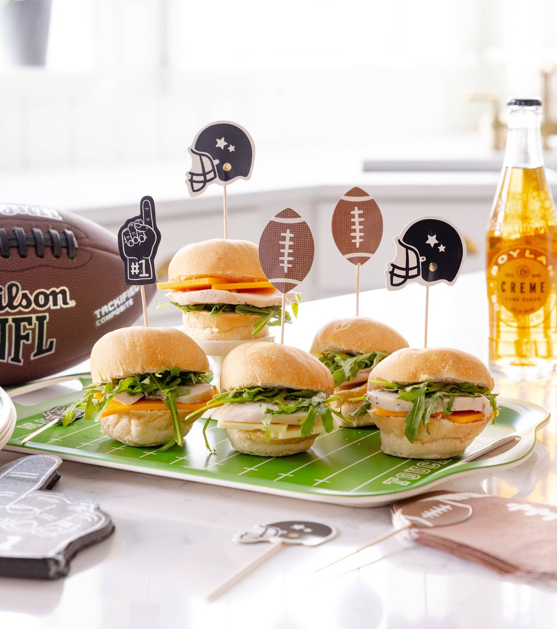 Mini burgers on a platter with football-themed picks, a drink bottle, and a football in the background.