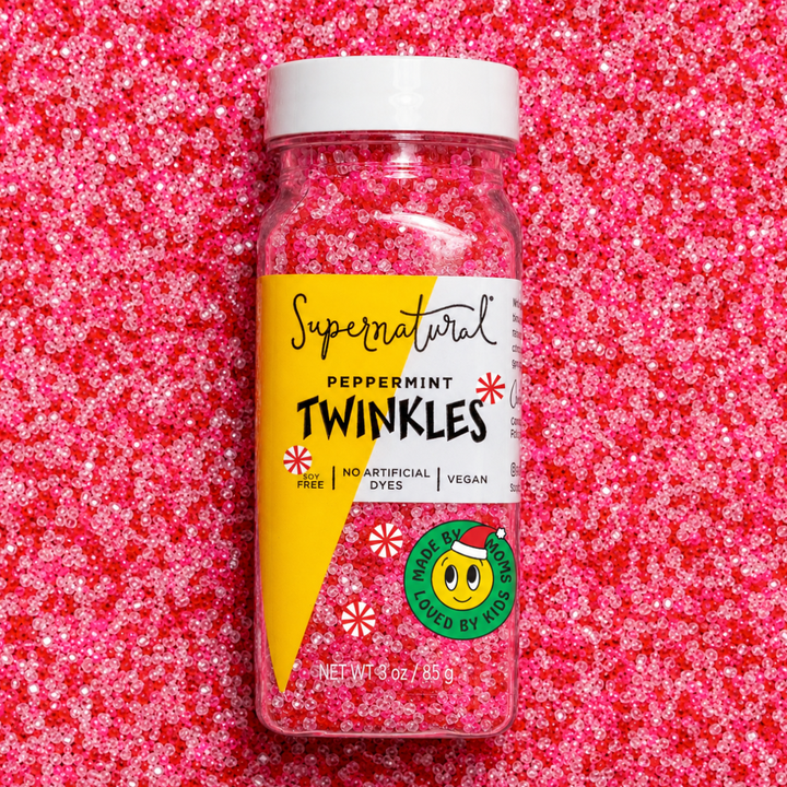 A clear plastic jar of Supernatural Peppermint Twinkles sprinkles with a white lid and a yellow-and-white label, placed on a textured pink sprinkles background.