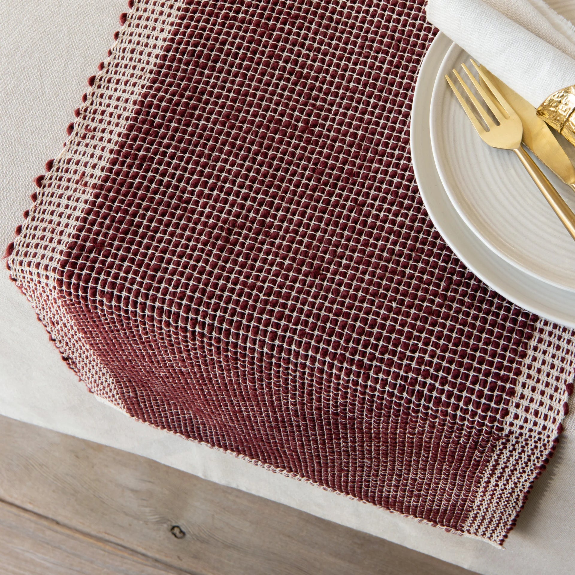 Woven table runner on a wooden table with a white plate and gold fork.