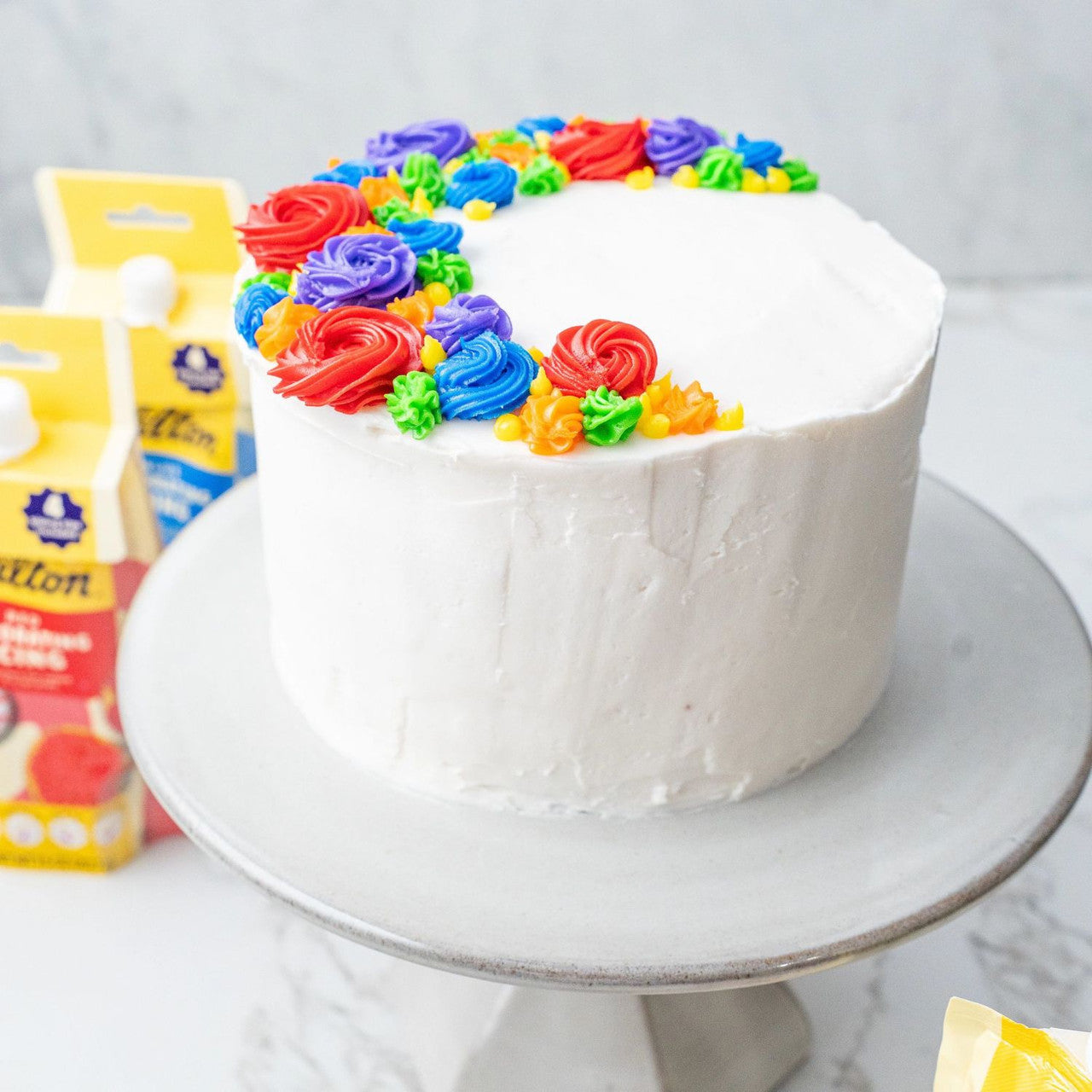 Colorful frosted cake on a white stand with frosting packets in the background.