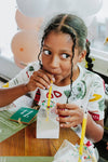 Child drinking from a milk carton with a straw at a table with balloons in the background