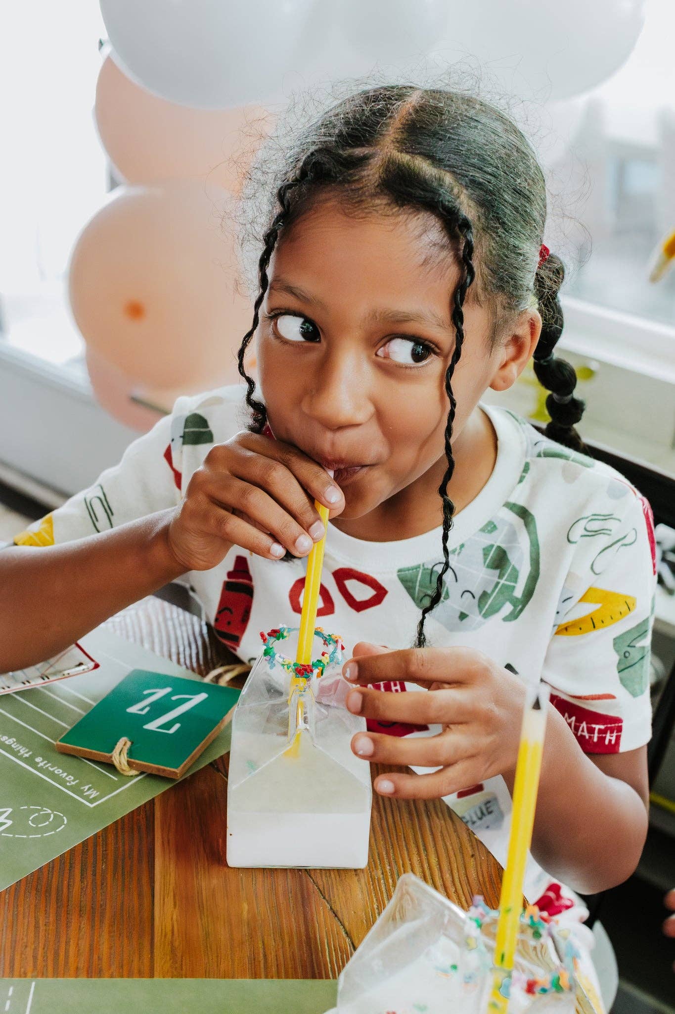 Child drinking from a milk carton with a straw at a table with balloons in the background
