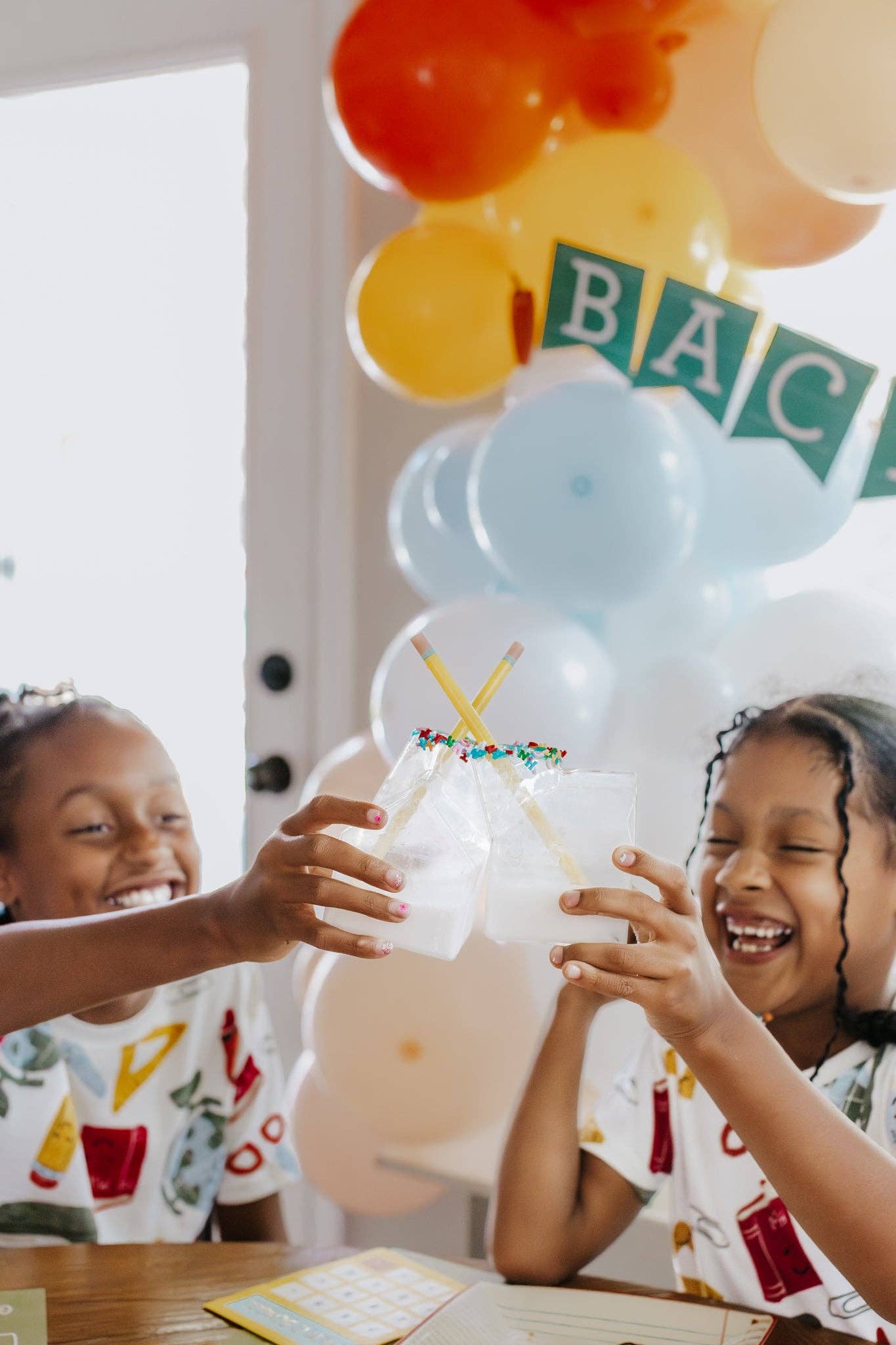 Two children celebrating with balloons and a cake, with 'BACK' letters visible.