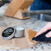 Person applying cutting board wax to a wooden cutting board with a tin of wax in the foreground.
