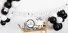 White and black Birthday decorations featuring banner, balloons, cups, plates, napkins, a birthday cake with topper on a white background