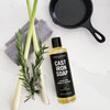 Bottle of Cast Iron Soap with a black cast iron skillet, green onions, and rosemary on a white surface.