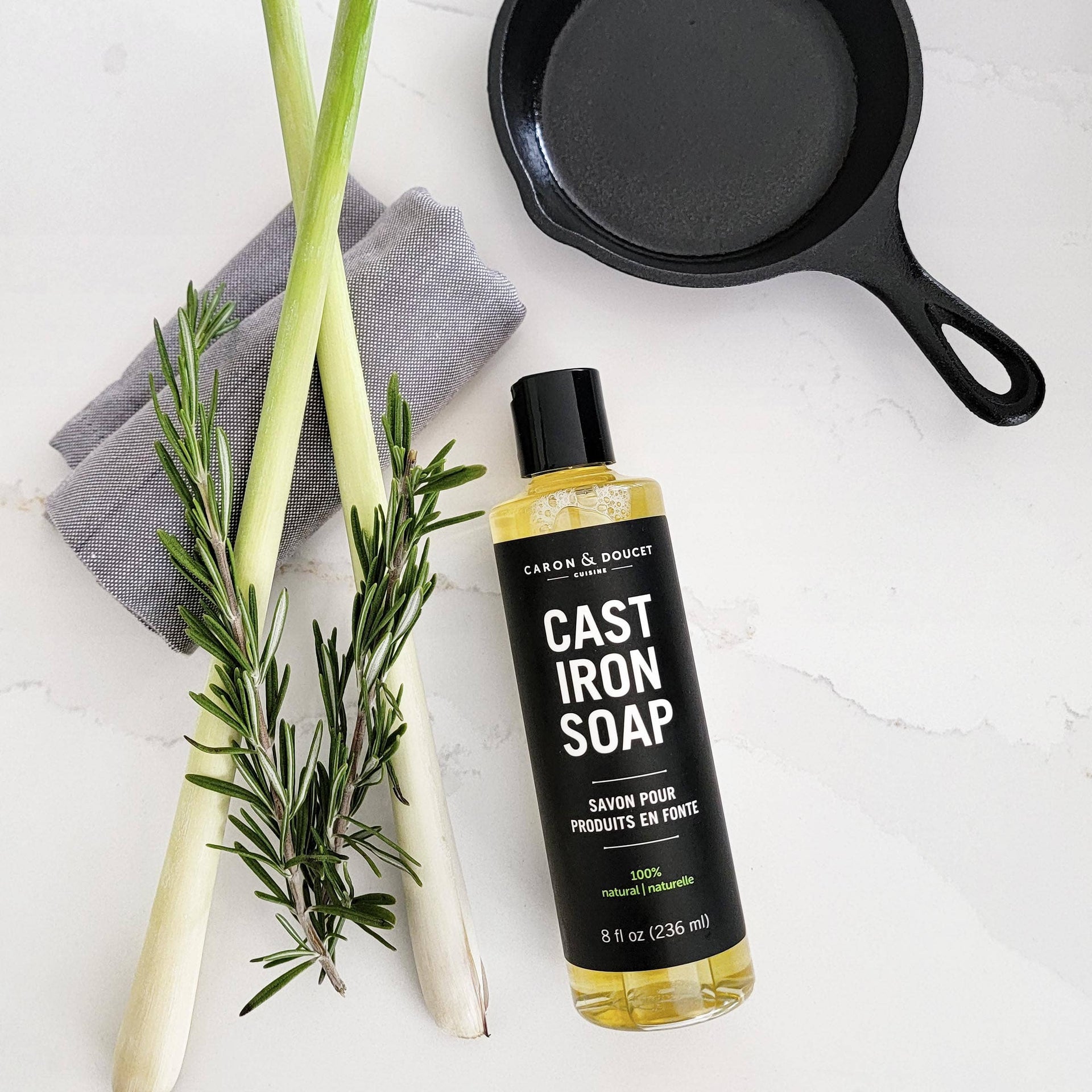 Bottle of Cast Iron Soap with a black cast iron skillet, green onions, and rosemary on a white surface.