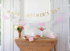 Decorative setup with 'Happy Mother's Day' banner, flowers, and a wooden table.
