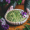 A baked asparagus and egg quiche tart in a white fluted pie dish, held by hands on a wooden table with purple flowers and asparagus scattered around.
