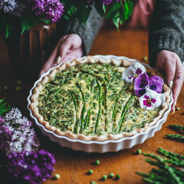 A baked asparagus and egg quiche tart in a white fluted pie dish, held by hands on a wooden table with purple flowers and asparagus scattered around.