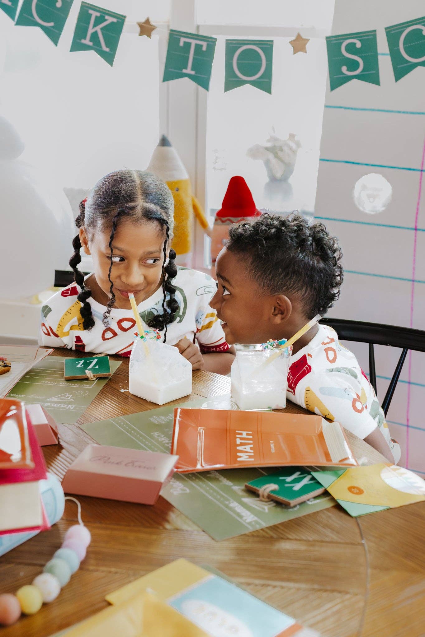 Two children sitting at a table with educational materials and a 'Back to School' banner in the background.