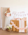 Woman in a long dress holding a pink plate next to a decorated table with 'It's a Girl' banner.