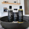Three bottles of Cast Iron Oil, Scrub, and Soap on a kitchen counter with a pan in the foreground.