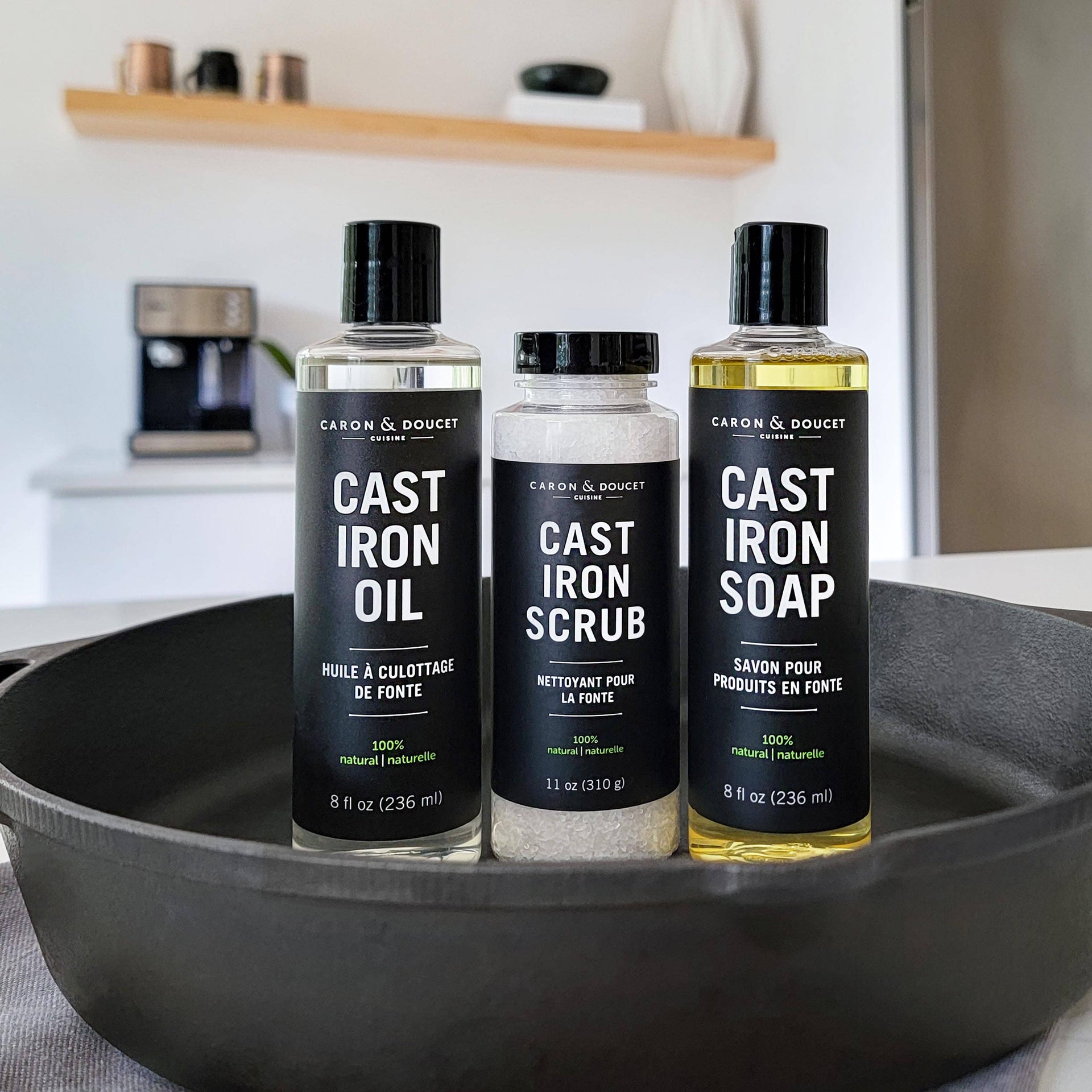 Three bottles of Cast Iron Oil, Scrub, and Soap on a kitchen counter with a pan in the foreground.