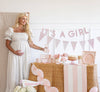 Pregnant woman in a white dress standing next to a 'It's a Girl' baby shower setup with pink decorations.