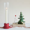 Taper candle holder with ribbed glass and red base sitting net to decorative Christmas trees against a white background.